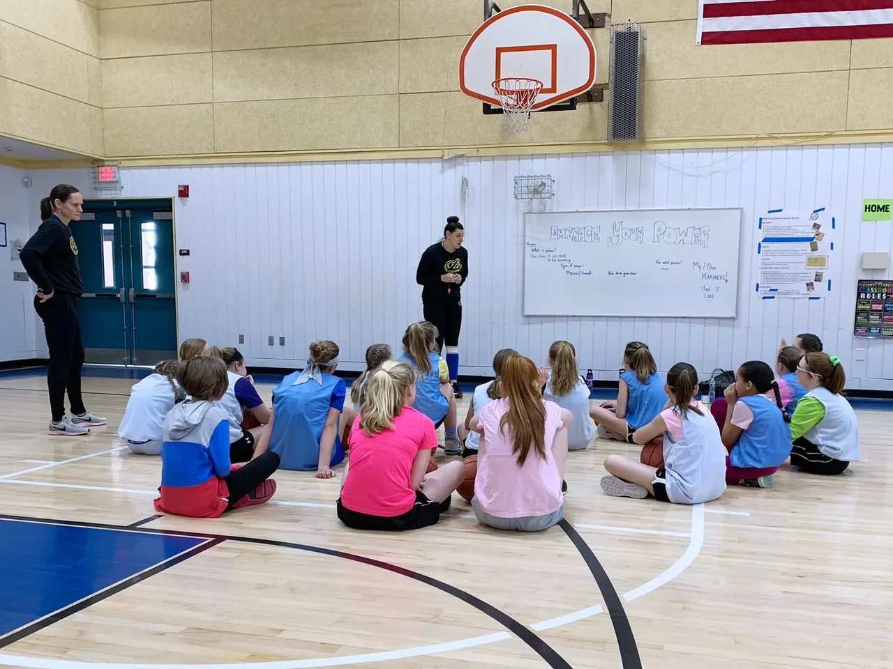 A clinic in a school gym: about twenty girls sit on the floor in a half circle facing two coaches, with EMBRACE YOUR POWER written on the whiteboard behind them.
