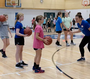Girls in a gym each holding a basketball at their hip, listening to a coach.