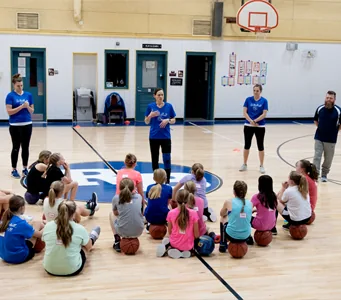 Players sit on the gym floor with their basketballs while coaches in blue shirts talk to them.