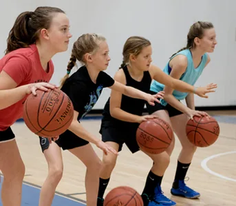 Four girls in a low stance, each holding a basketball out in front, working on a drill side by side.