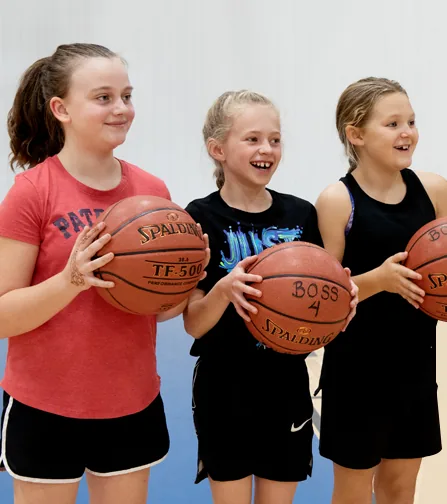 Three smiling girls standing in a gym, each holding a basketball; one ball has BOSS 4 written on it.